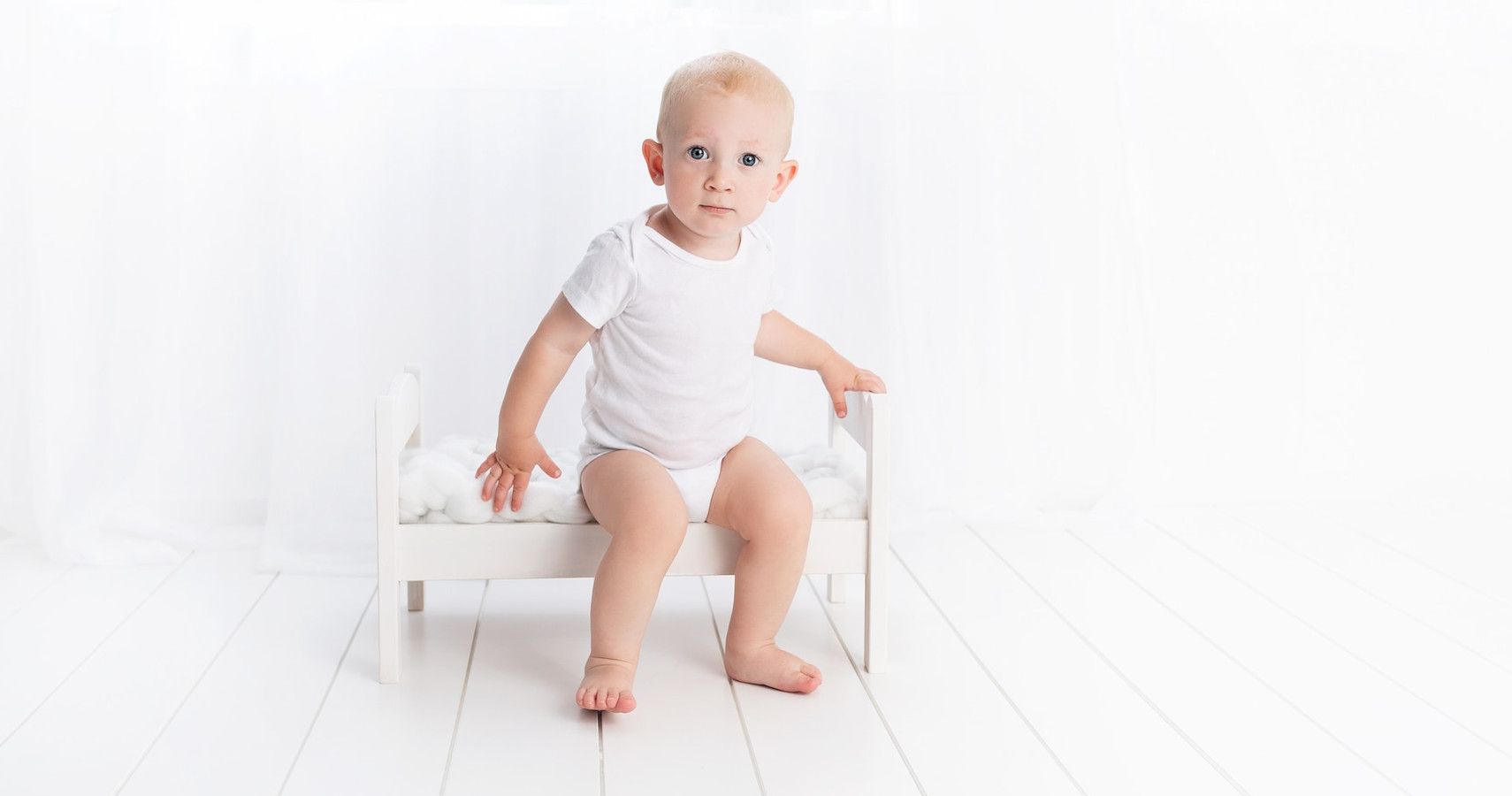 A baby in white sitting on a white bed on a white background.