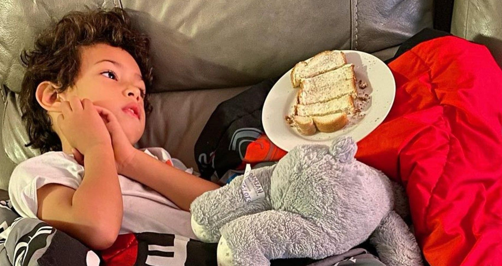 A child laying down on the couch with a plate of food that he is barely eating